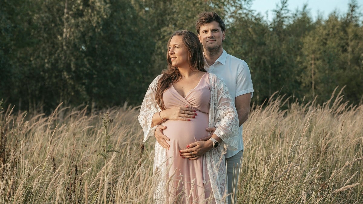 An expectant couple standing together in a field of tall summer grass at golden hour