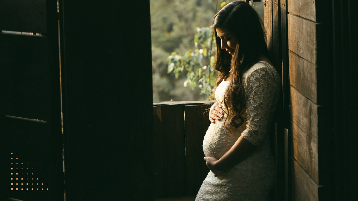 A pregnant woman in a white lace dress stands at an open wooden door, looking down at her bump